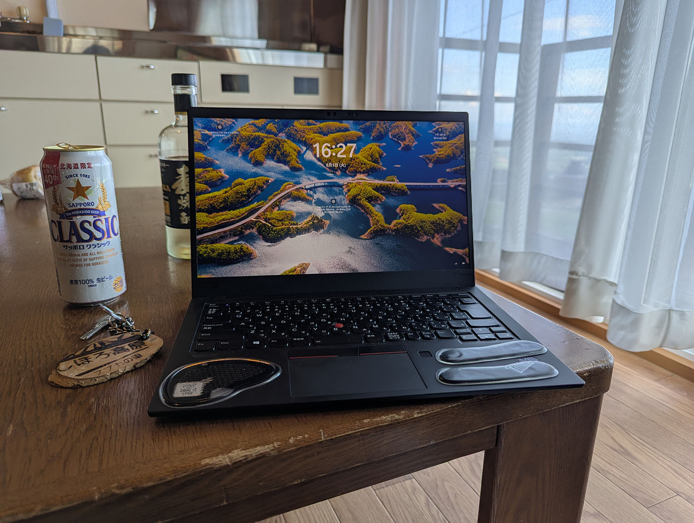 ロッジの一室でノートパソコンを開いた様子。机の上にはサッポロクラシックの缶ビールと焼酎の瓶、宿泊用の木札キーが置かれ、窓からは柔らかな日差しが差し込んでいる。