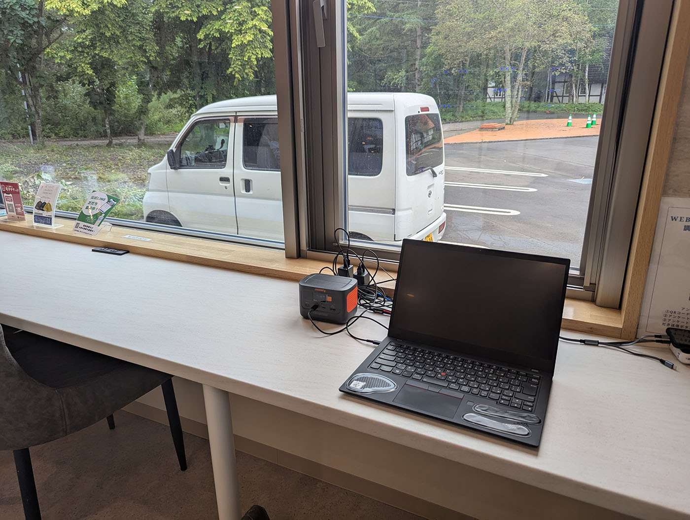 十勝エアポートスパそらの館内、窓際のカウンター席でノートPCを広げてノマドワークをする様子。窓の外には駐車場に停めた車が見える。