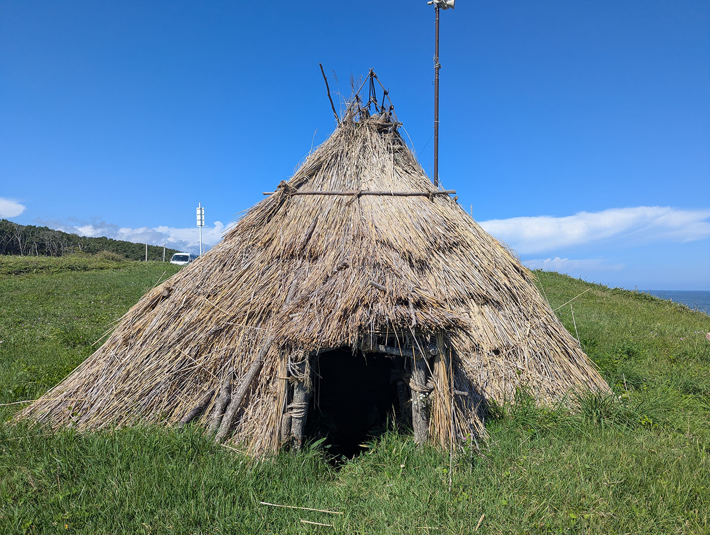 ホロカヤントー竪穴群の復元竪穴住居。茅で覆われた円錐形の屋根が青空の下に立ち、去年よりも整備が進んでいる様子がうかがえる。