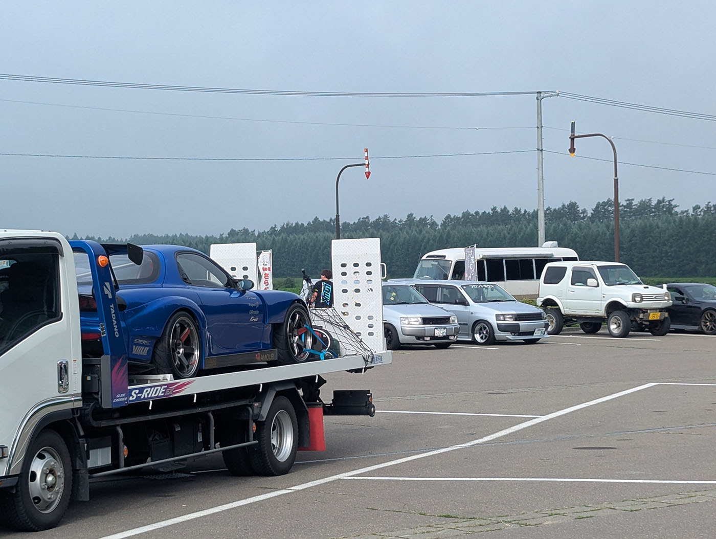 道の駅の駐車場に並ぶカスタムカーやスポーツカー。積載車に載せられた青い車や改造車が目を引く。