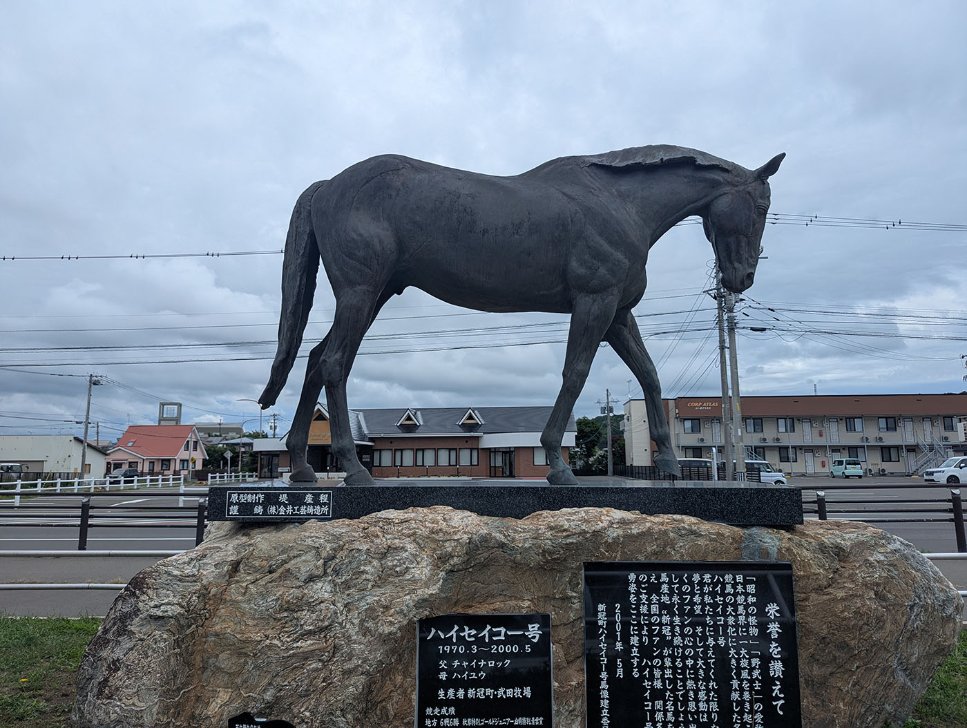 優駿・ハイセイコー号の銅像。下部の石碑には経歴や功績が刻まれている。背景には牧場風の建物や道路、電線が広がる。