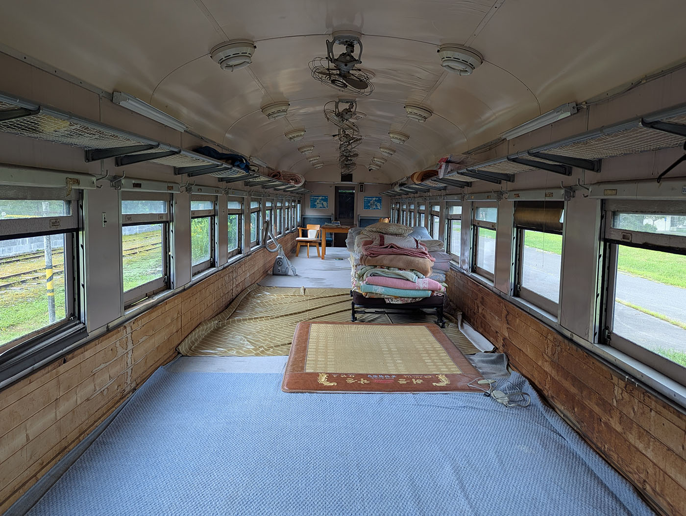 北海道むかわ町の富内銀河ステーションに保存された青い客車の内部。窓が並ぶ車内には畳とカーペットが敷かれ、布団や座卓が置かれた宿泊用スペースとして活用されている。