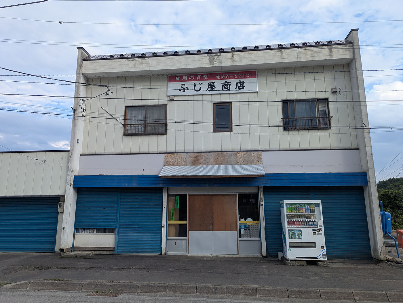 北海道むかわ町の富内銀河ステーション利用時に料金を支払う「ふじ屋商店」。青いシャッターと白い外壁が特徴で、店先には自動販売機が置かれている。