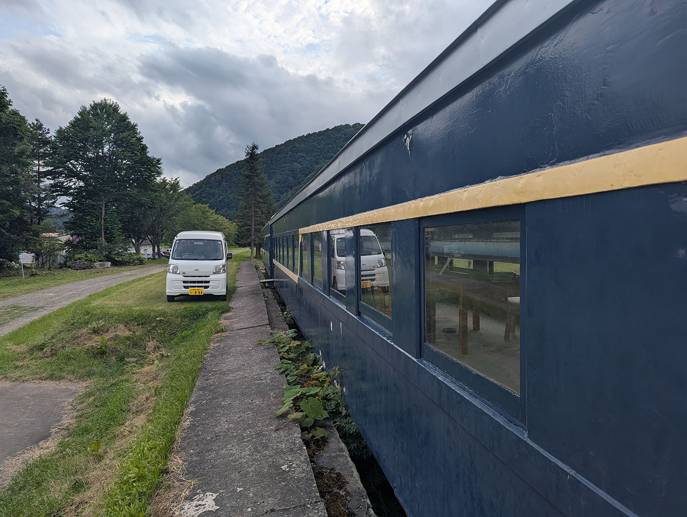 北海道むかわ町の富内銀河ステーションに保存された青い客車。背後には緑豊かな山々が広がり、車両横の芝生には白い軽バンが停まっている。
