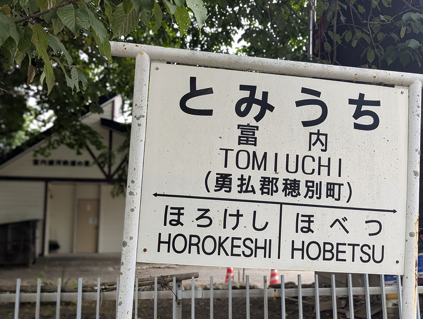 富内駅の駅名標。「とみうち」「TOMIUCHI」と表記され、左に「ほろけし」、右に「ほべつ」の案内がある。背景には木立と駅舎が見える。