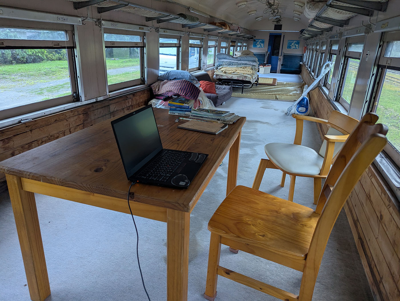 古い客車を改装した車内に置かれた木製テーブルと椅子。テーブルにはノートパソコンが開かれ、窓の外には緑の景色が広がる。奥には布団や座椅子、掃除機などが置かれ、生活感が漂う。