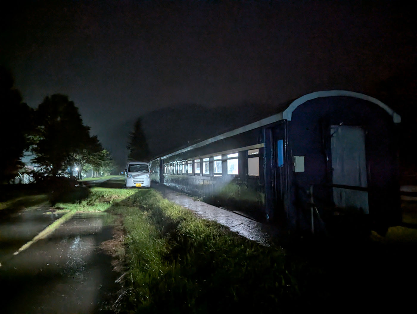 夜の闇の中、車内に灯りがともった古い客車が静かに停まっている。外は雨で、列車の窓明かりだけが周囲を柔らかく照らしている。