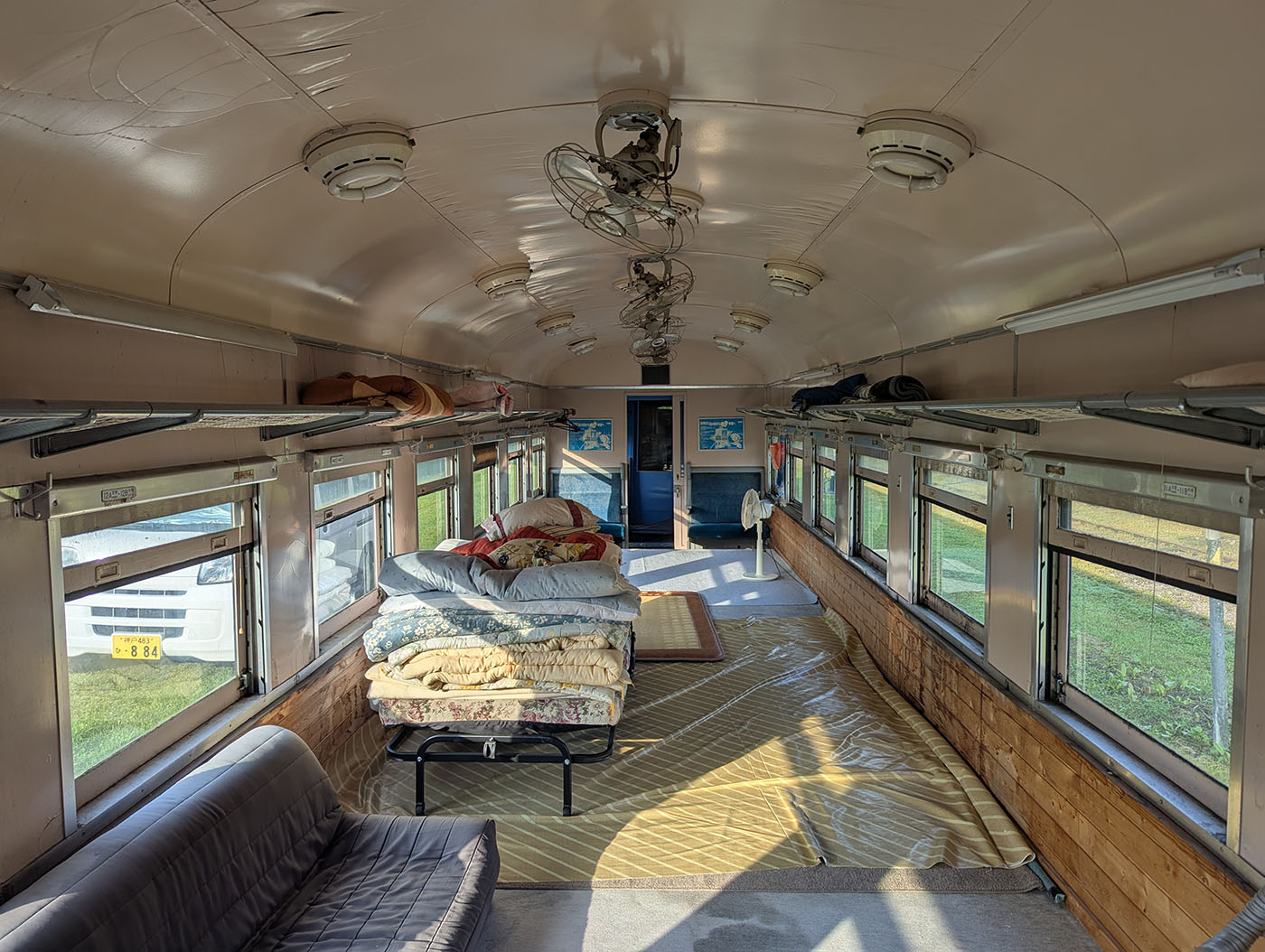 富内銀河ステーションに保存された青い客車の車内。窓際には布団が積まれ、ソファや扇風機が置かれた簡素な宿泊スペースが広がっている。天井には扇風機や照明が並び、当時の雰囲気を残している。