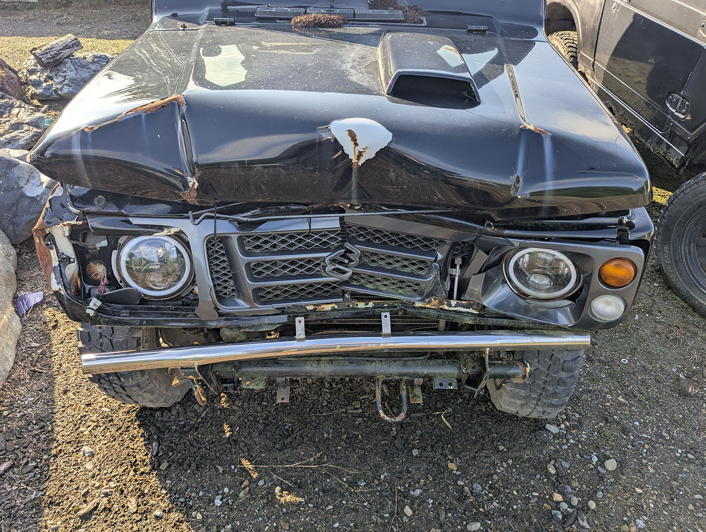 正面から見た黒いジムニー。左右のライトやバンパーに損傷が残る。