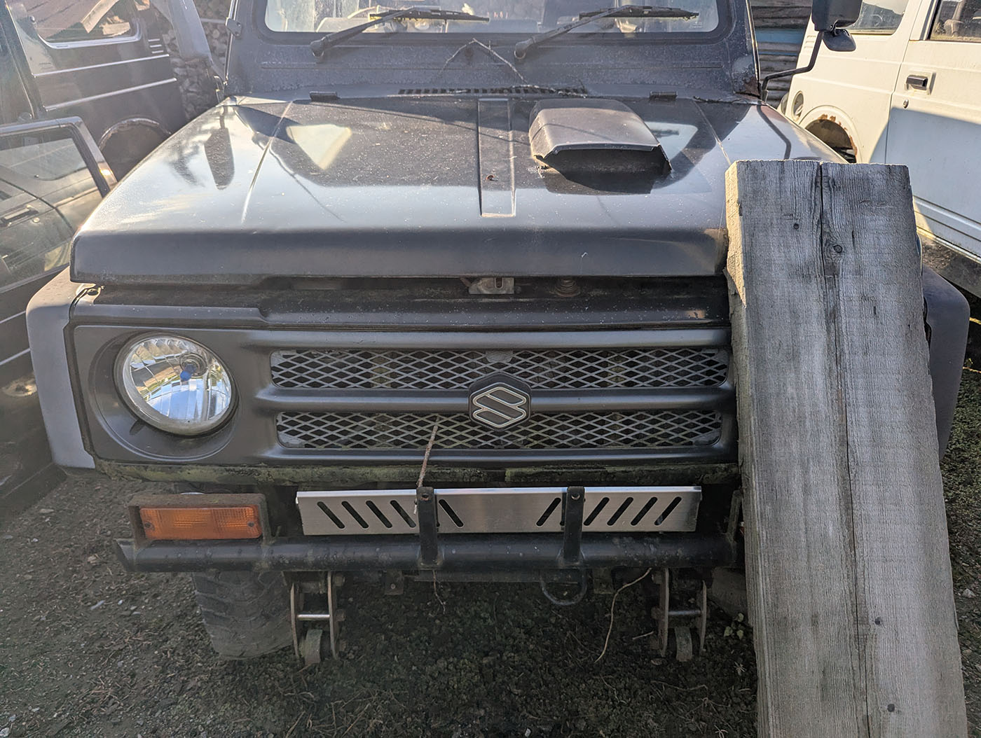 黒いジムニーの正面アップ。グリル中央にはスズキのエンブレム。右側に木の板が立てかけられている。