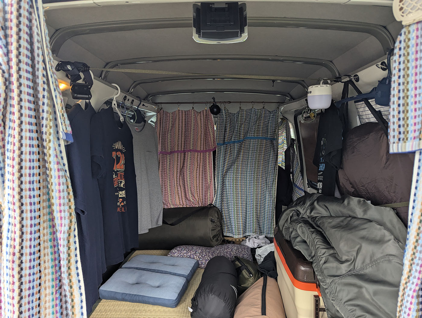 軽バンの車内を仕切りカーテンや衣類で覆い、長期駐車や車中泊に備えた遮光対策の様子。寝袋やクッション、収納ボックスが並び、生活空間として整えられている。