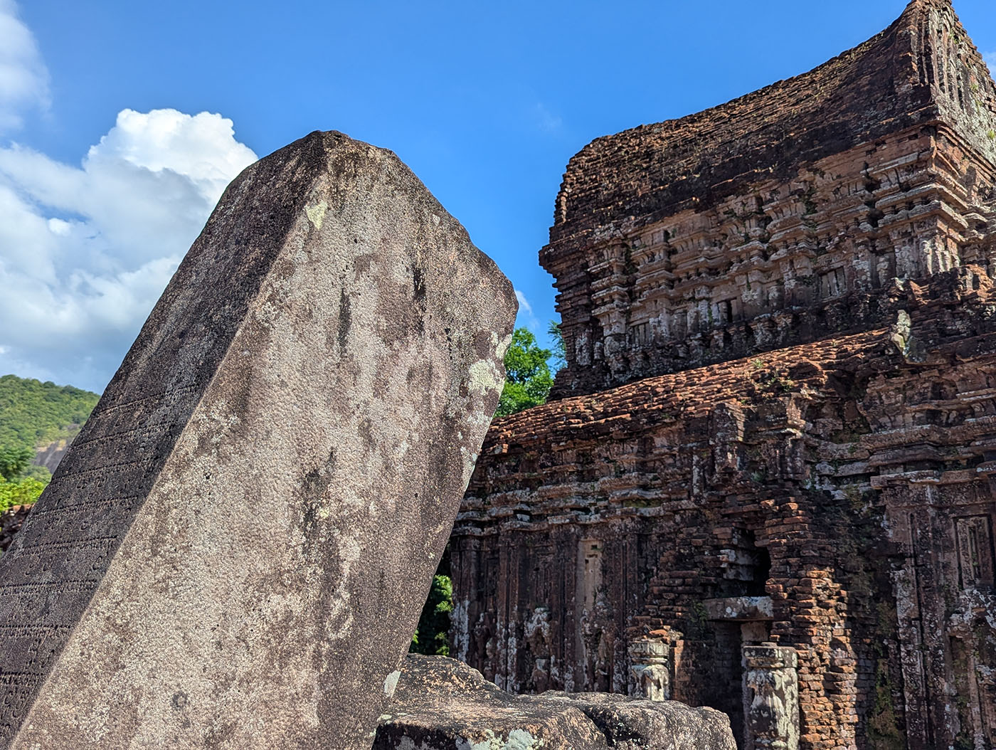 倒れた石碑に刻まれた古代文字と、その奥にそびえるミーソン遺跡のレンガ造りの塔