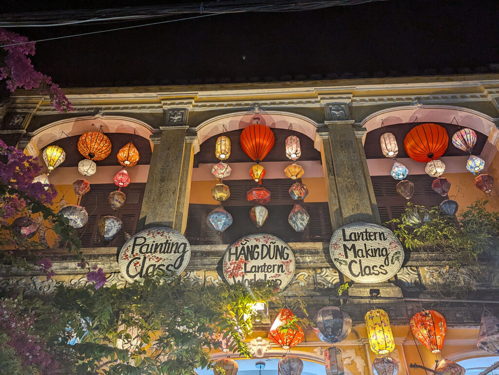ホイアン旧市街の二階建て建物に吊るされたカラフルなランタンと、ランタン作りやペインティングの看板