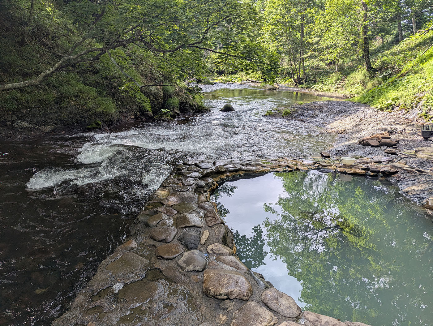 ユウヤンベツ川の流れのすぐ隣に湧く鹿の湯温泉。石で囲まれた湯船に木々の緑が映り込む、自然と一体になれる秘湯。