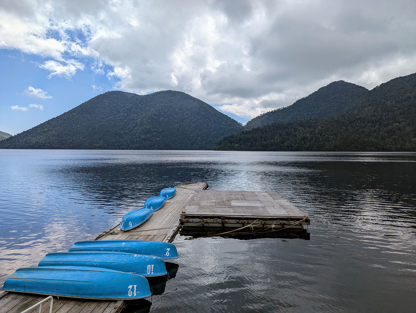 然別湖の桟橋に並ぶ青い手漕ぎボート。山々が湖を囲み、曇り空が静かに広がる湖面に映っている。
