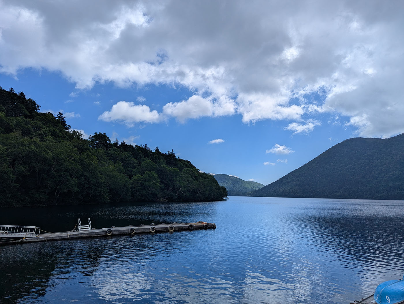 然別湖の桟橋と湖面の風景。青空と雲が映り込む静かな湖に、山々の稜線が包み込むように広がっている。