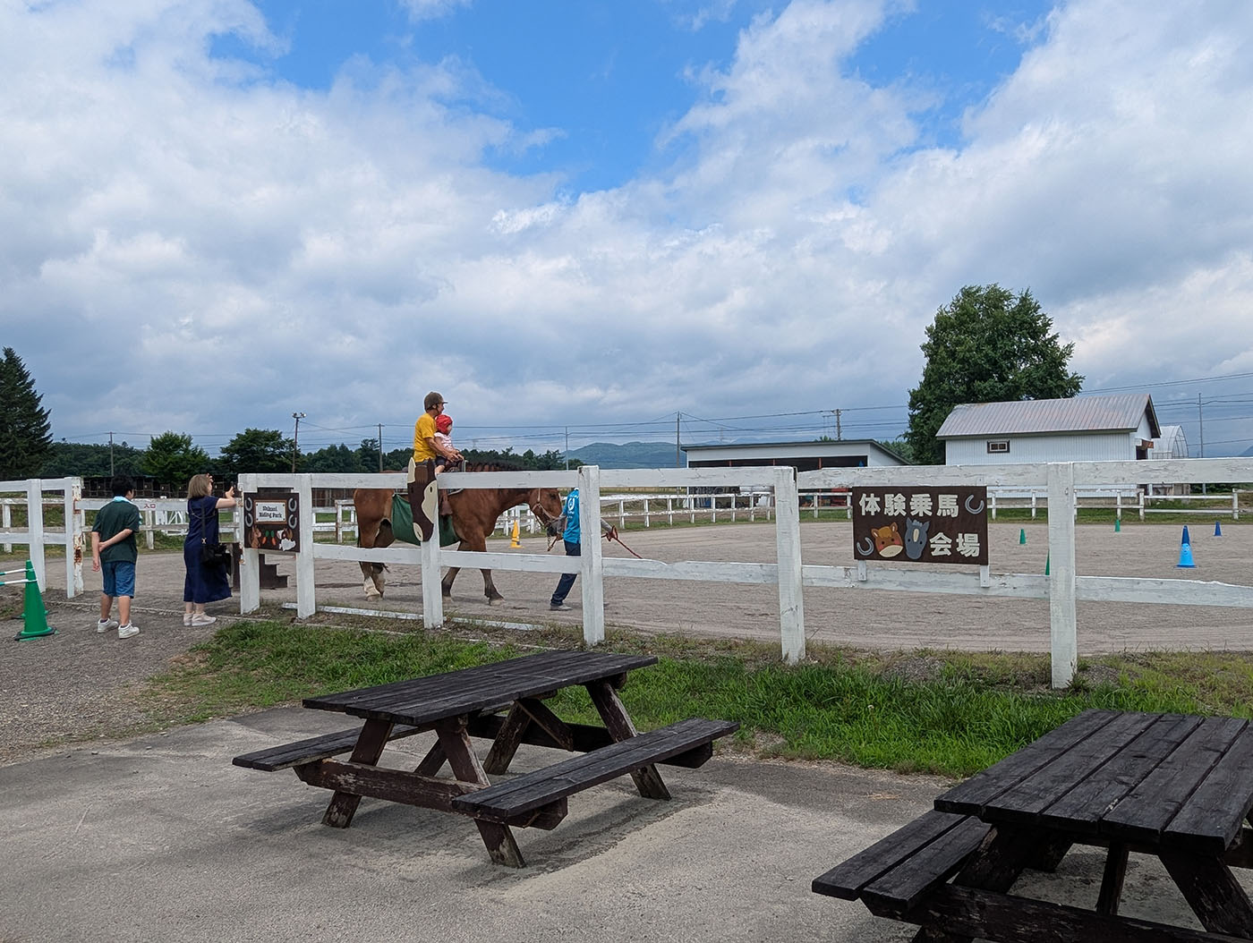 体験乗馬会場で馬に乗る親子と、見守るスタッフや家族