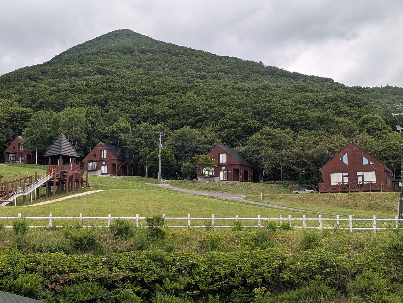ヌプカの里のコテージ群と大型木製遊具、背景には緑の山と森林が広がる。