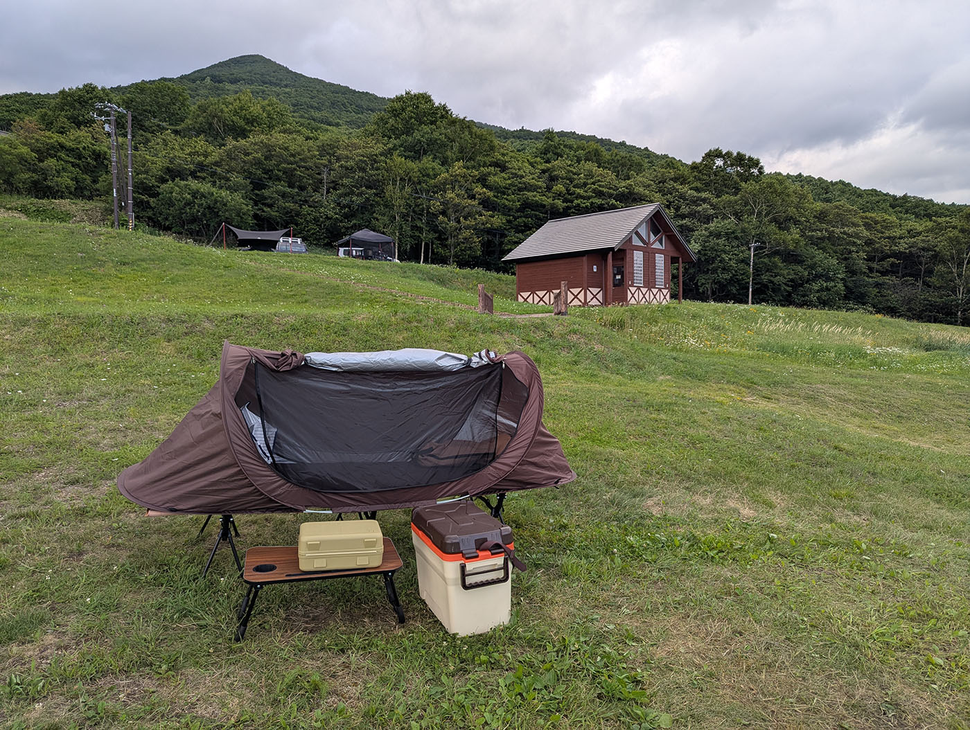 ヌプカの里のキャンプサイトに設営されたコットテントとテーブル、クーラーボックス。背景にはロッジと緑の山並みが広がる。