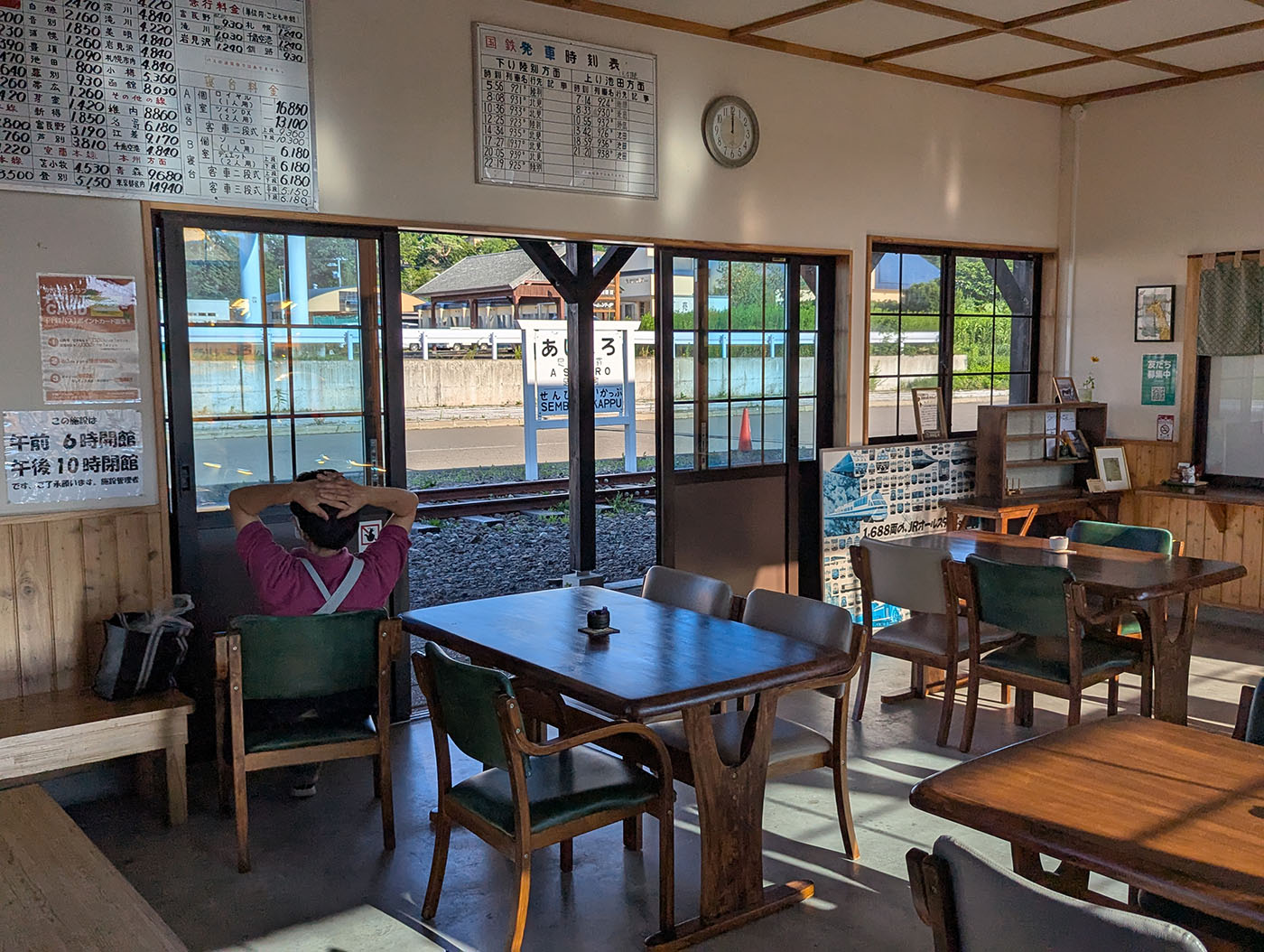 旧足寄駅舎を利用した待合室の内部。木製テーブルと椅子が並び、窓の外には線路と「あしょろ駅」の駅名標が見える。ベンチにはご年配の女性が線路の方を向いて座っている。