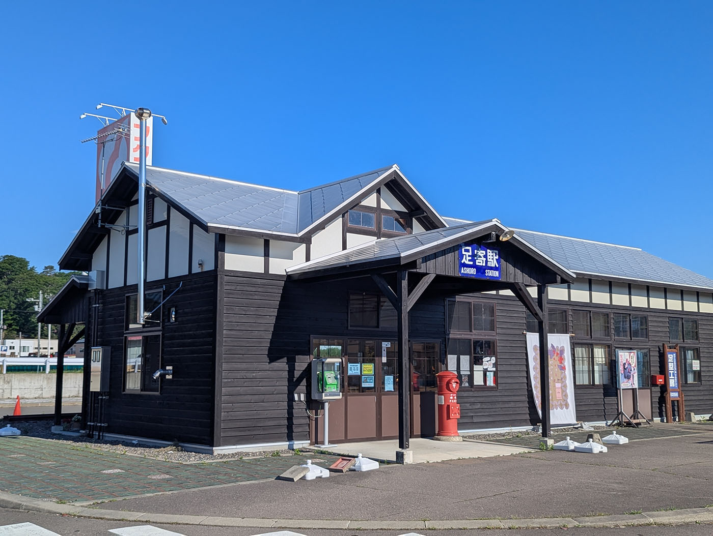澄み渡る青空の下に建つ旧足寄駅舎の全景。黒い木造の外壁に「足寄駅 ASHORO STATION」の駅名看板が掲げられ、正面には赤い丸型ポストが置かれている。