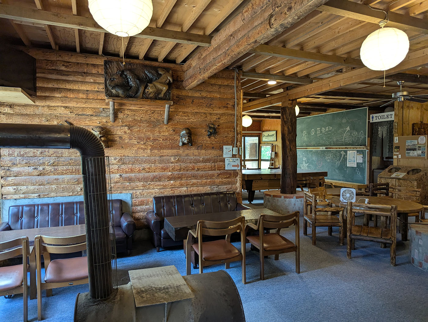 木のぬくもりに包まれたクリオネキャンプ場のラウンジ。壁には木彫りの装飾や面が飾られ、薪ストーブが中央に配置されている。