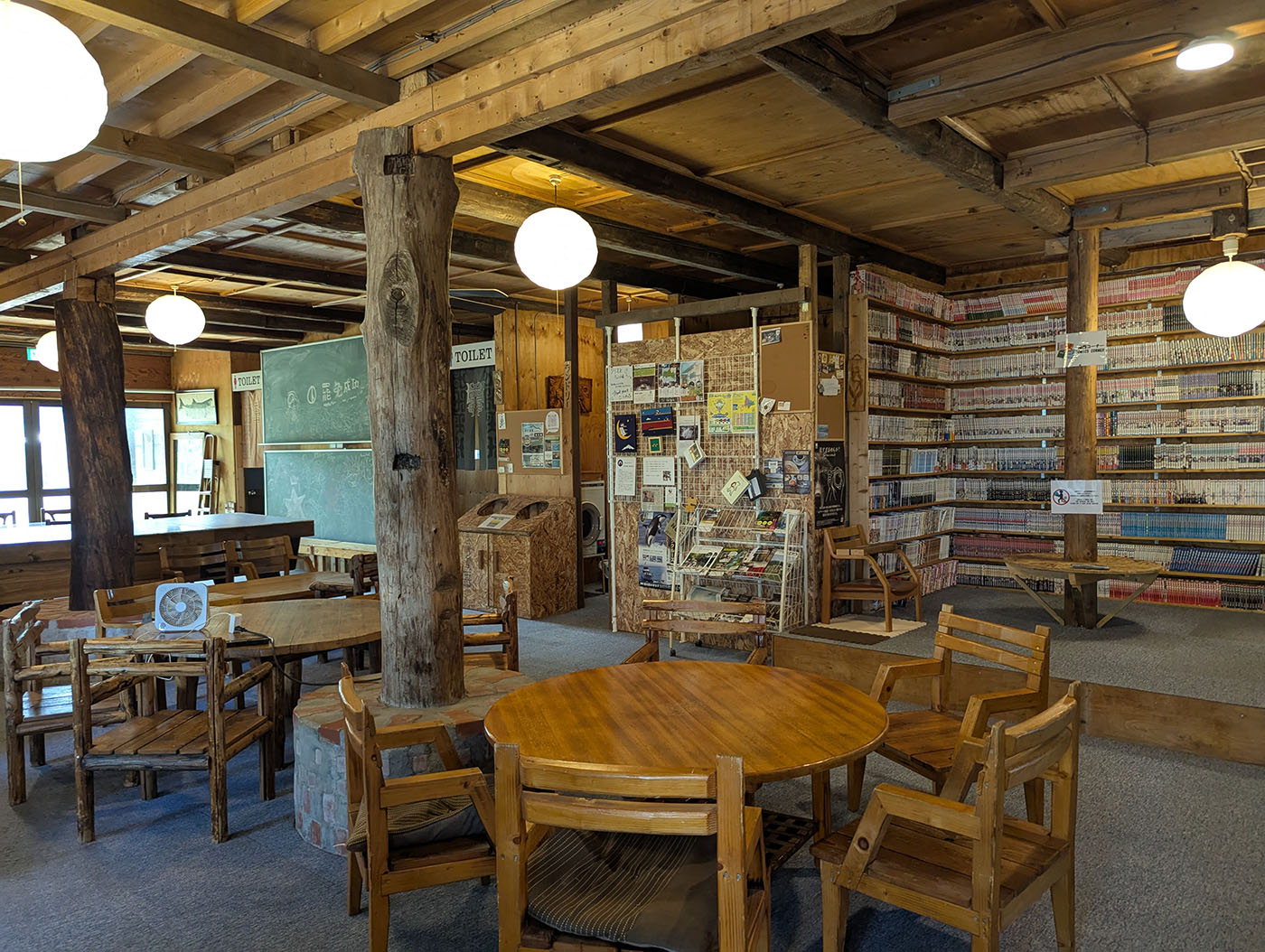 クリオネキャンプ場のログハウス管理棟のラウンジスペース。木製の丸テーブルと無数の漫画が並ぶ壁一面の本棚が特徴的。