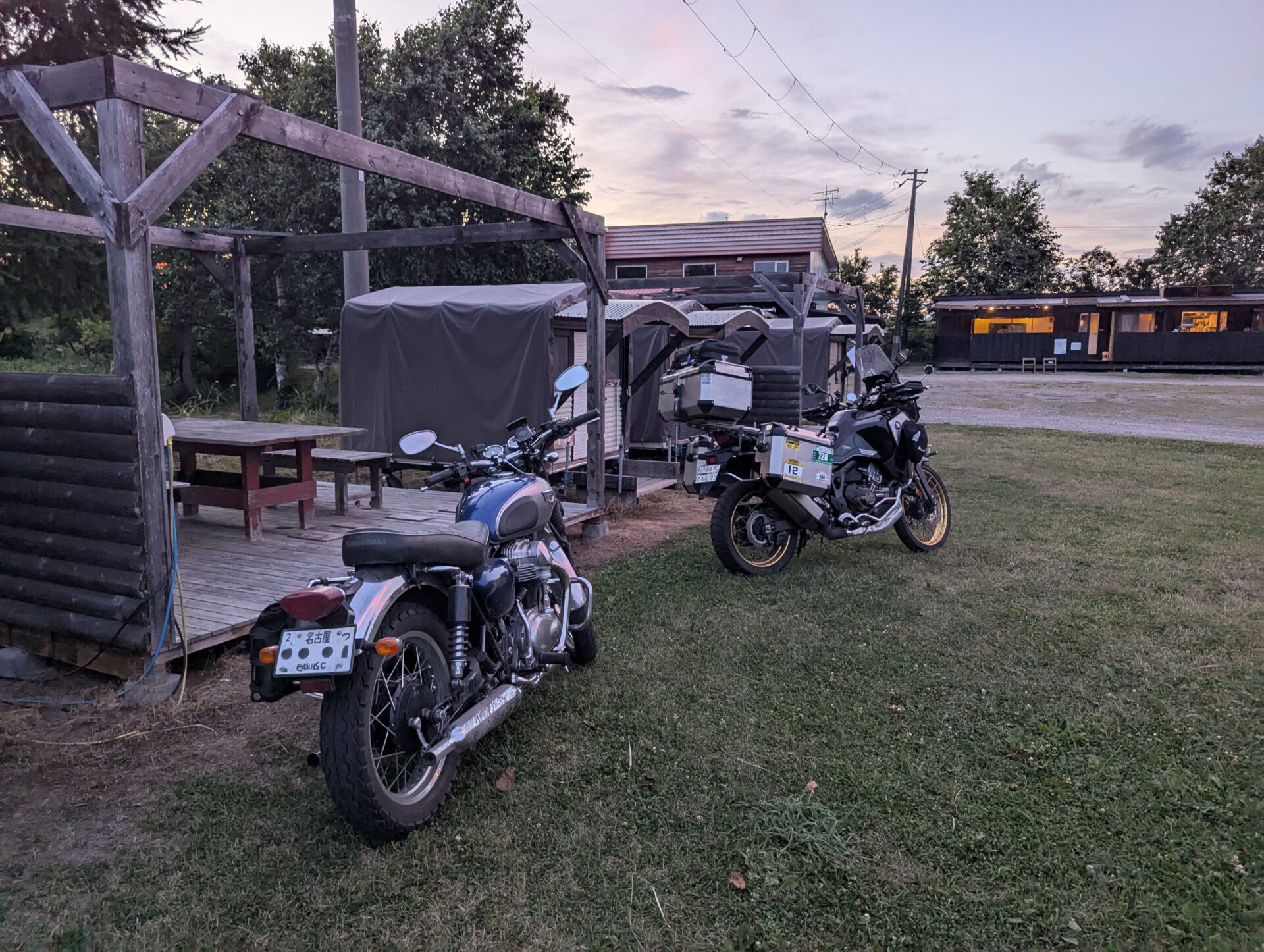 夕暮れのクリオネキャンプ場に停車する2台のバイクと、背後に並ぶモバイルハウス。ライダーたちの旅の拠点としての雰囲気が漂う。