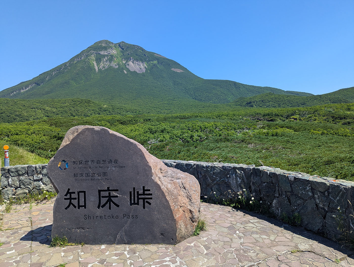 知床峠の展望スポットに立つ石碑と、その背後にそびえる羅臼岳。青空と緑の山肌が広がる。