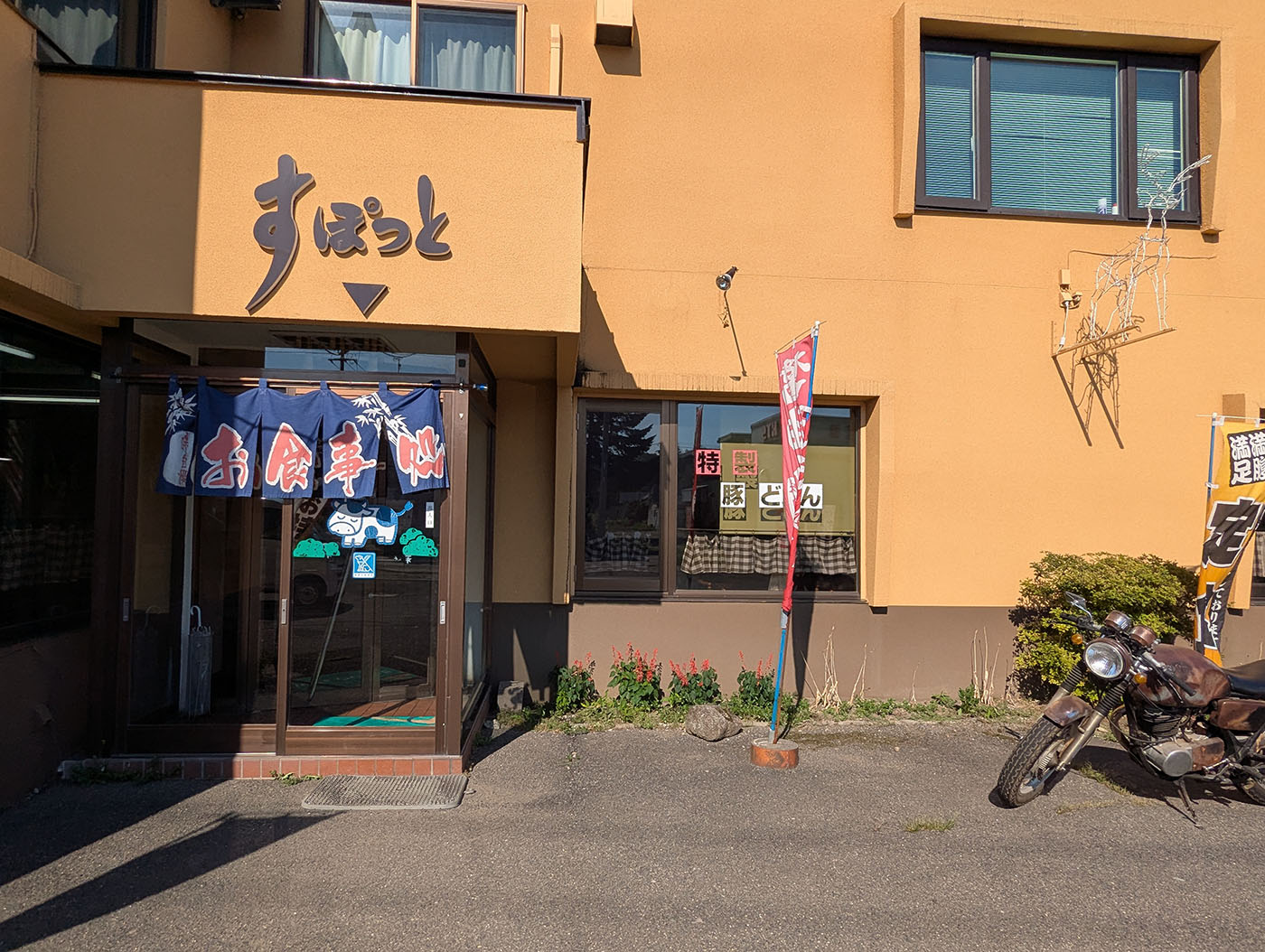 鹿追町のレストラン「すぽっと」の店舗外観。青い暖簾と「豚どん」の文字、店先には年季の入ったバイクが停められている。