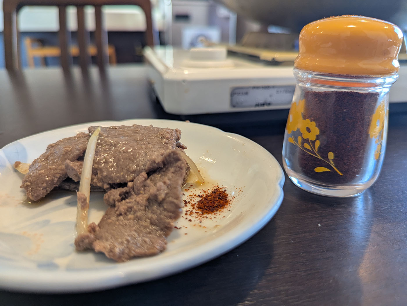 鹿追町「大阪屋」の煮込みジンギスカンを小皿に盛りつけ、一味唐辛子と共に提供された様子。奥にはレトロな黄色いフタのスパイスボトルが置かれている。