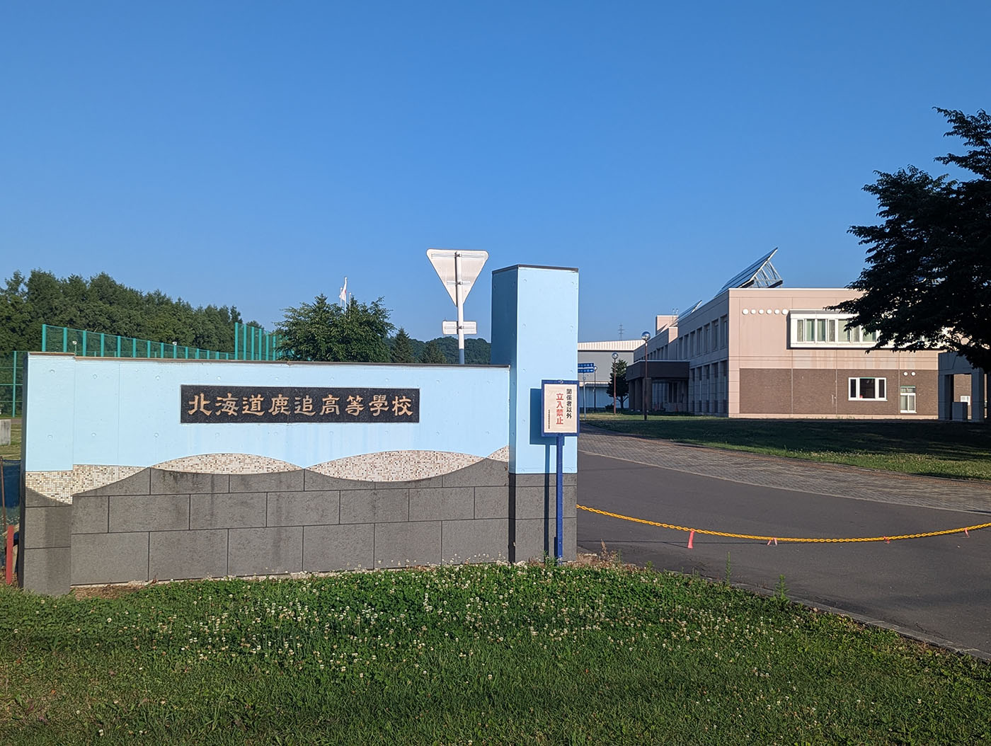 北海道鹿追高等学校の正門と校舎。校門には校名が掲げられ、奥には太陽光パネルを備えた校舎が見える。青空の下に立つモダンな校舎が印象的。