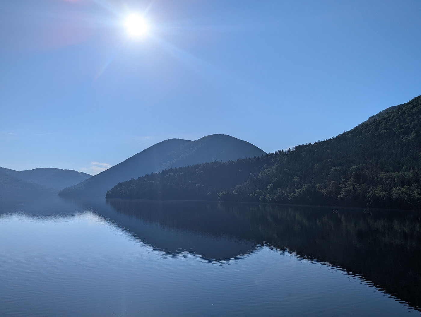 静かな然別湖の湖面に映る山々と、朝日に照らされた澄んだ青空の風景