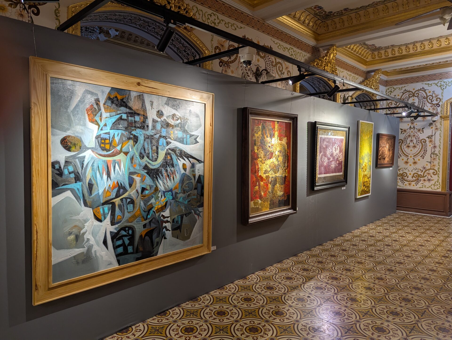 金色の装飾が施された豪華な室内で、灰色の展示壁に現代アートや伝統的絵画が並ぶ美術展の一角。手前には抽象的な大作、奥には金箔や細密画が並んでいる。