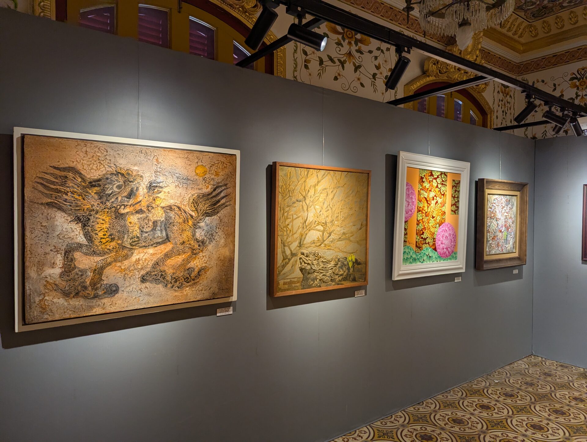 灰色の展示壁に沿って、麒麟のような神獣を描いた茶系の絵画、枯れ木と石を描いた静謐な作品、鮮やかな花と緑を描いた色彩豊かな作品、細密なパターンアートが並ぶ美術展の一角。