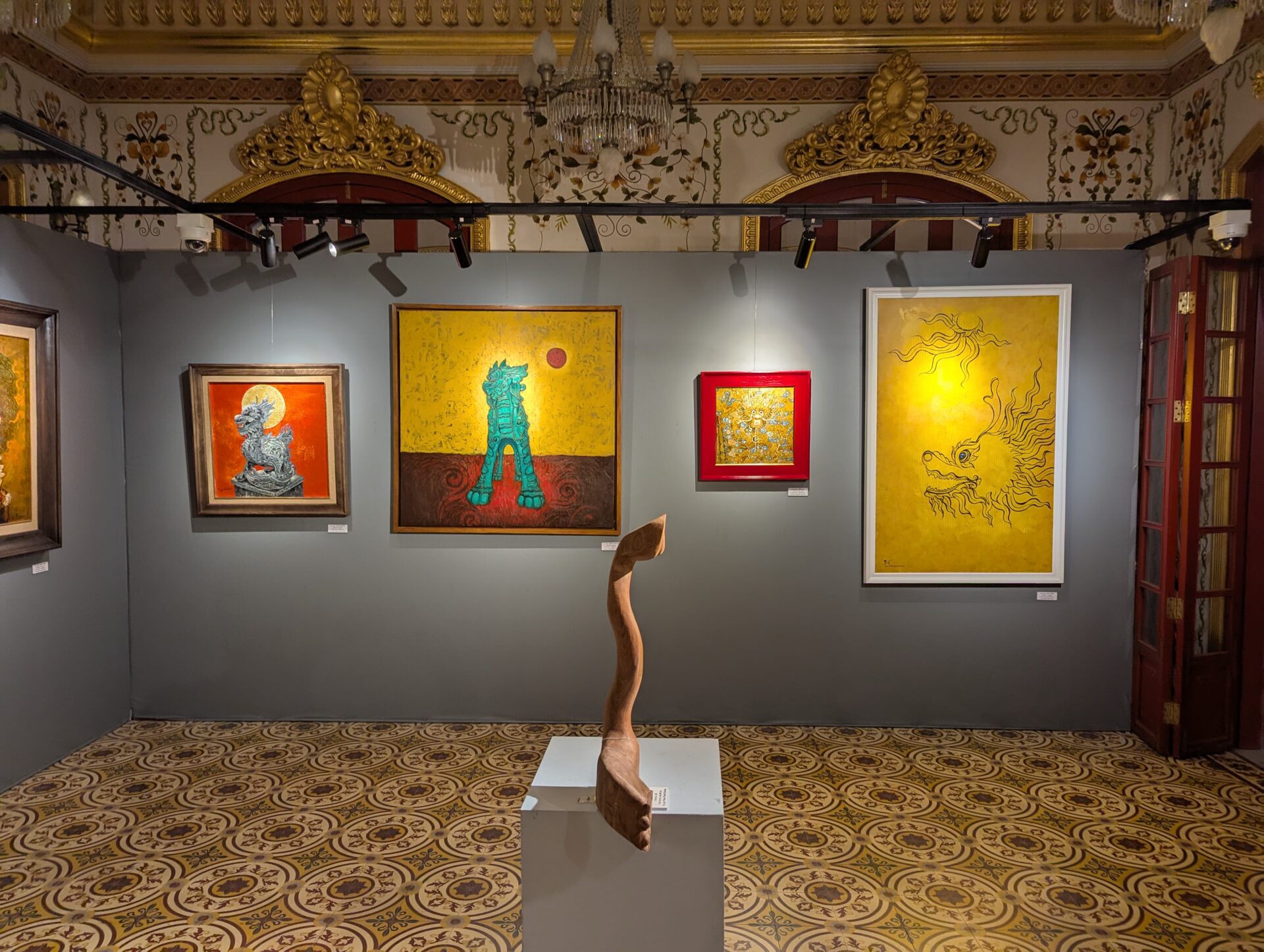 灰色の展示壁に、麒麟や龍をモチーフにした鮮やかな絵画が並び、中央には木製の抽象彫刻が置かれた美術展の一角。背景には黄色や赤を基調とした作品が映える。