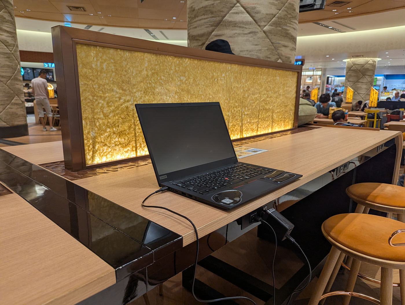 伊丹空港の充電カウンター席でThinkPadを使用している様子。木目調のカウンターテーブルに黒いノートPCが置かれ、背景には金色に光る仕切りパネルと空港のターミナル内が見える