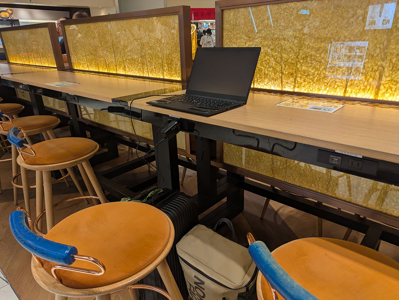 伊丹空港充電カウンターの全体像。木目調の長いカウンターテーブルにThinkPadが置かれ、オレンジ色の椅子が並んでいる。テーブル下には電源コンセントやUSBポートが完備されている