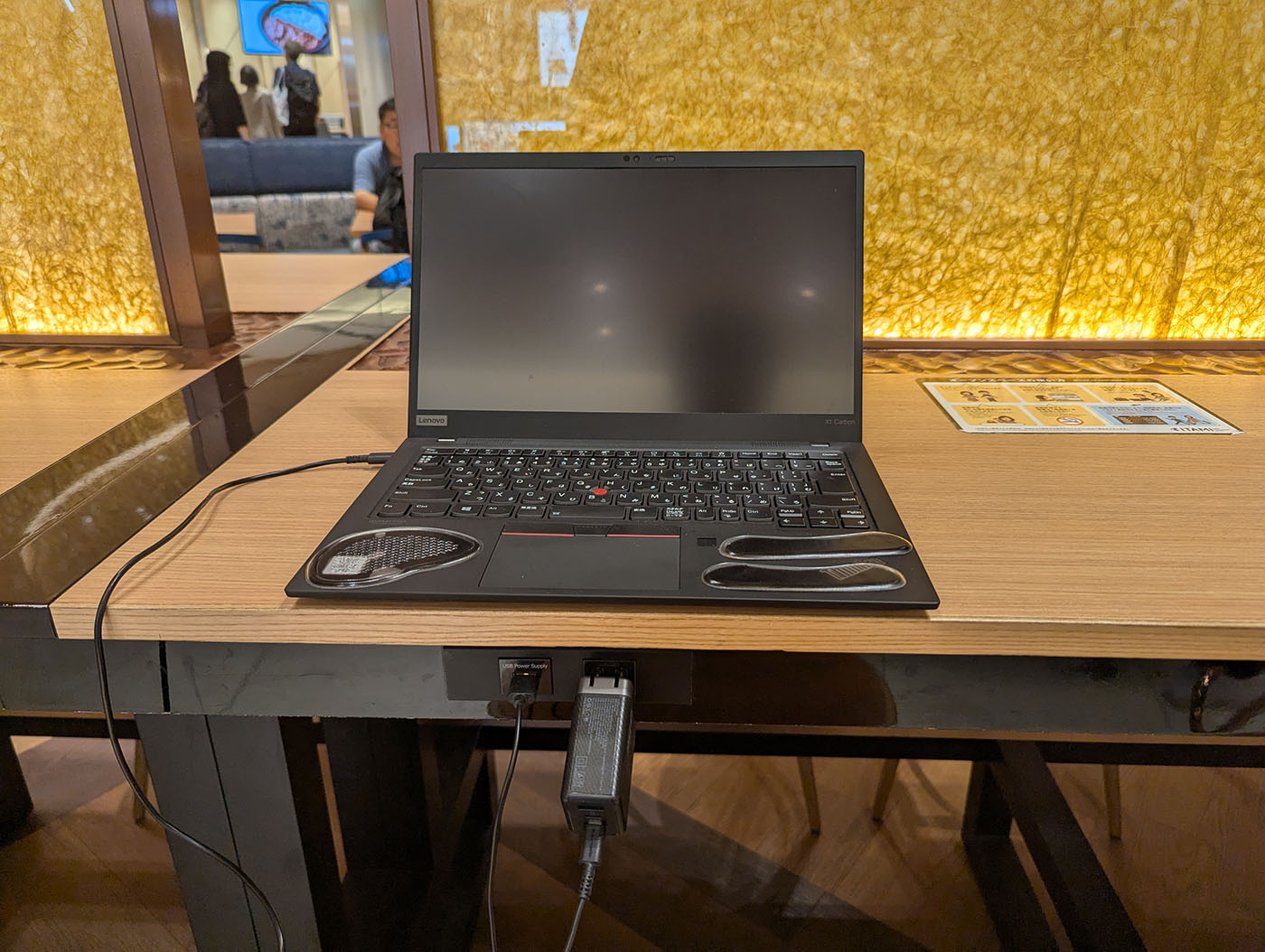 ThinkPadのクローズアップ。木目調テーブルの上に置かれた黒いノートPCで、電源アダプターがテーブル下のコンセントに接続されている様子が見える