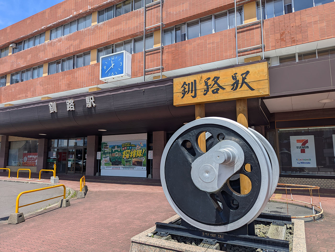 釧路駅正面と、駅前に設置された巨大な車輪モニュメント。青空に映える駅舎とシンボル。