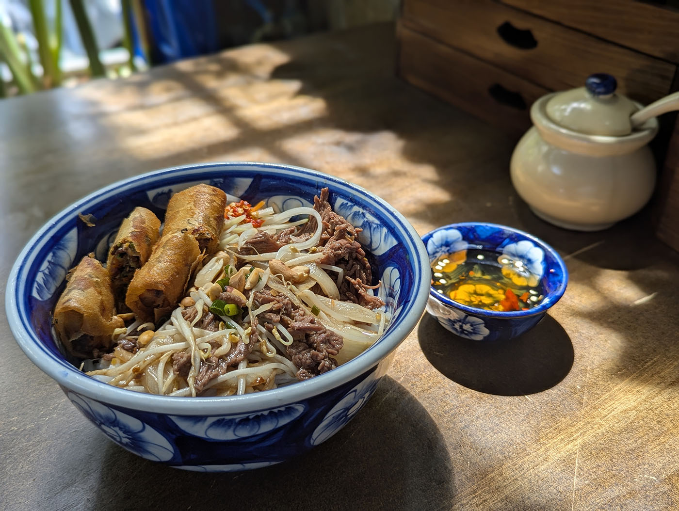 青い陶器の器に盛られた牛肉と揚げ春巻き入りのフォーと、横に添えられたヌクチャムのタレ