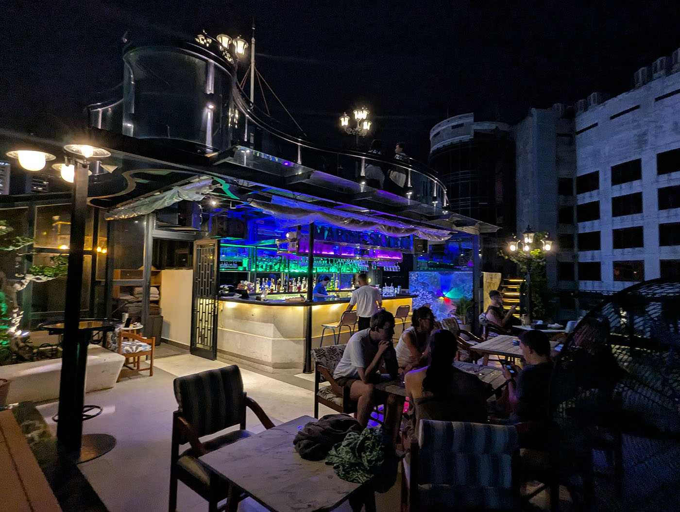 夜の屋上バー。青と紫のライトに照らされたカウンターと、テラス席でくつろぐ人々。
