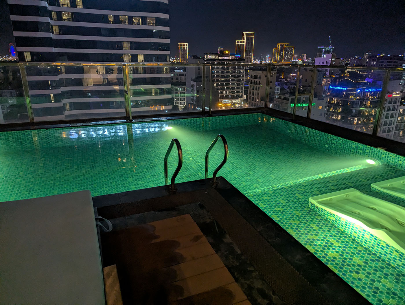 夜の屋上プール。水中照明に照らされて緑がかった水面が輝き、背後にはダナン市街の夜景が広がる。