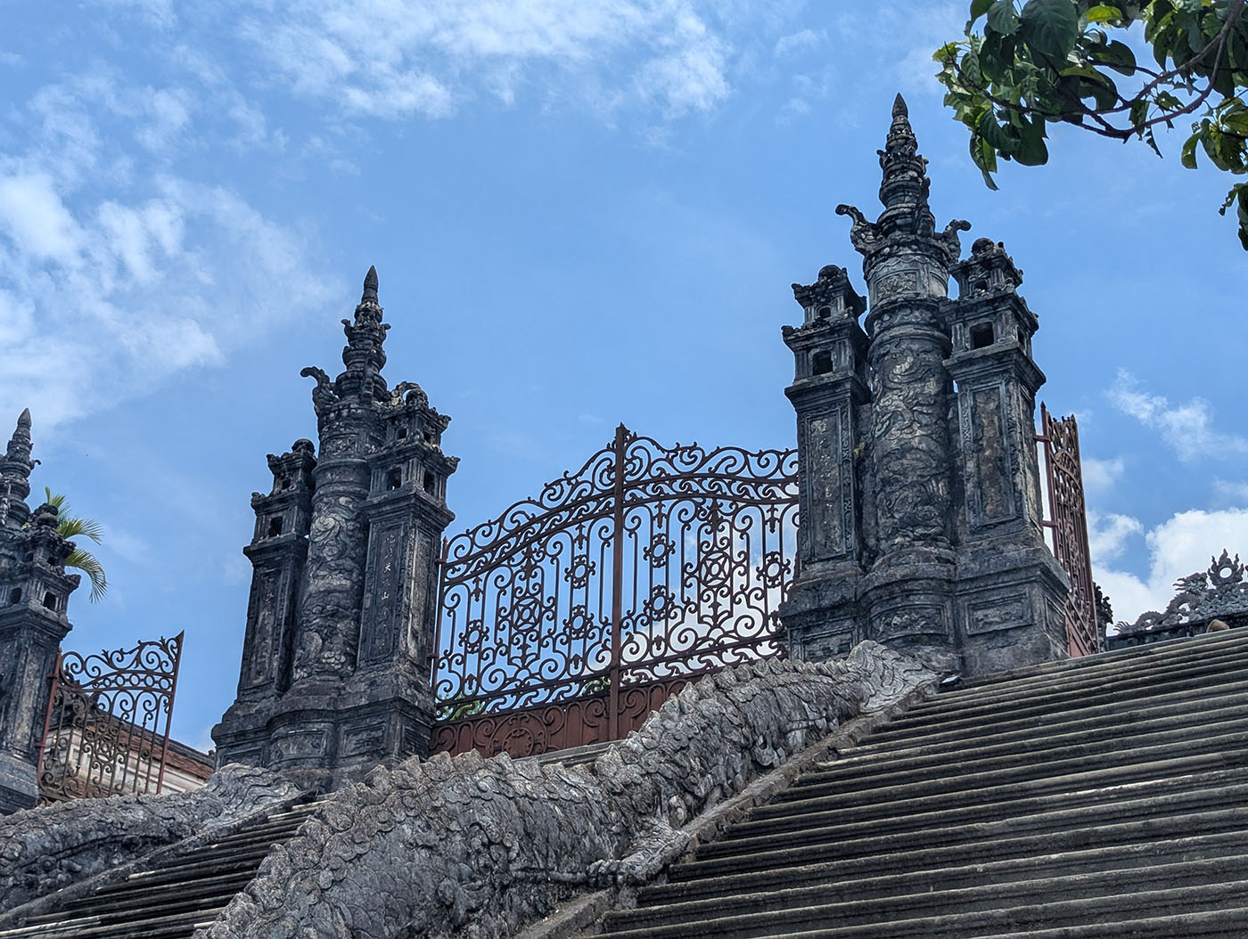 石造りの門と階段を見上げた構図。精巧な装飾が施された塔のような門柱がそびえ立ち、青空が広がる。