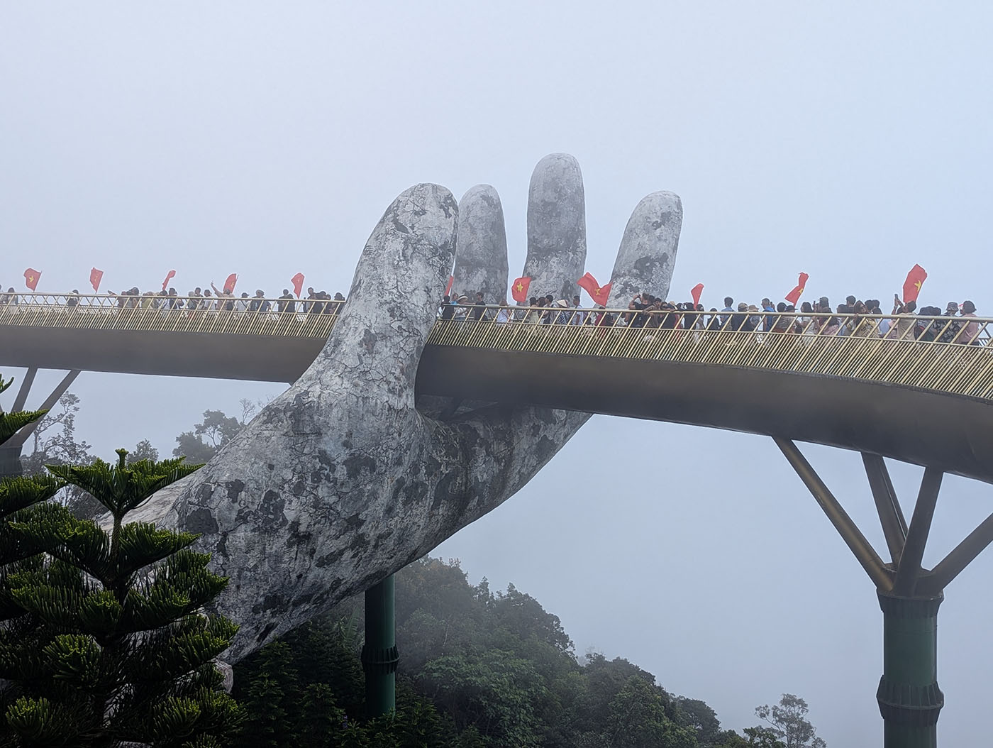 霧に包まれた巨大な石の手に支えられたゴールデンブリッジを、多くの観光客とベトナム国旗が彩る