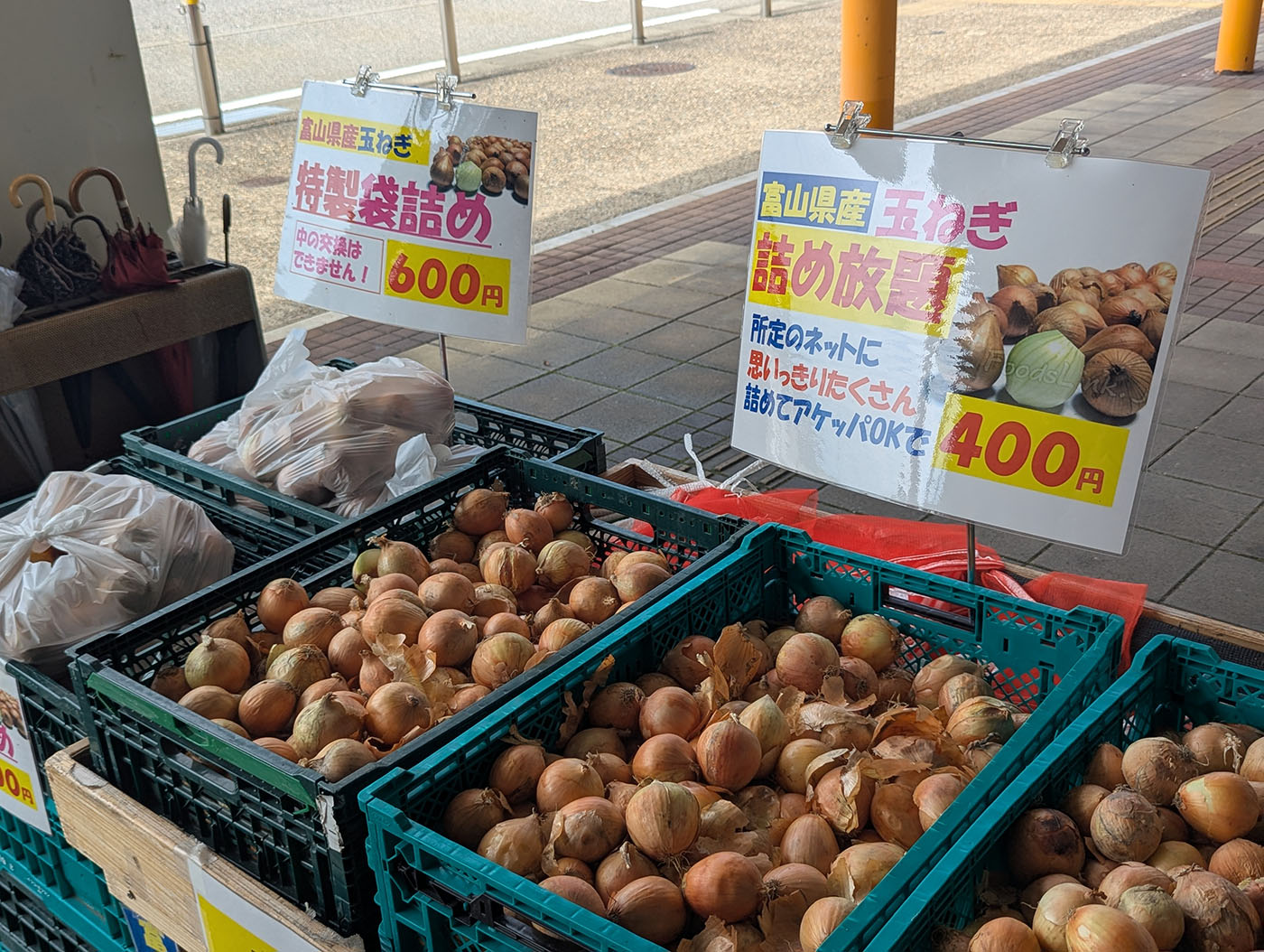 道の駅メルヘンおやべの入口に並ぶ玉ねぎの直売コーナー。「富山県産玉ねぎ詰め放題400円」「特製袋詰め600円」の看板とともに、黒いコンテナに山盛りの玉ねぎが並ぶ。