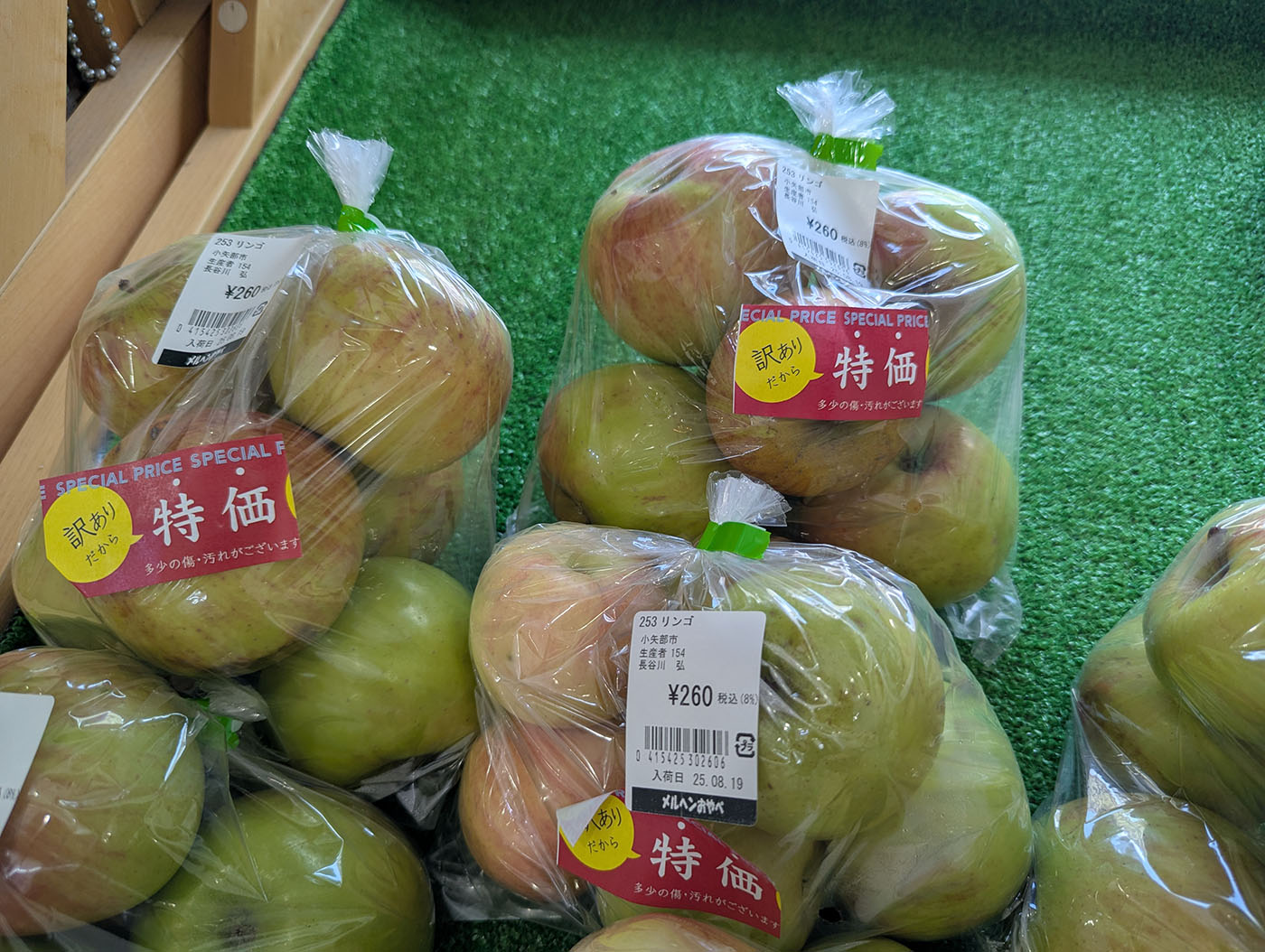 緑色の人工芝の上に並べられた袋入りリンゴ。「訳あり 特価」のシールが貼られ、260円の価格表示がある。ふじりんごが3〜4個ずつ透明な袋に詰められている。