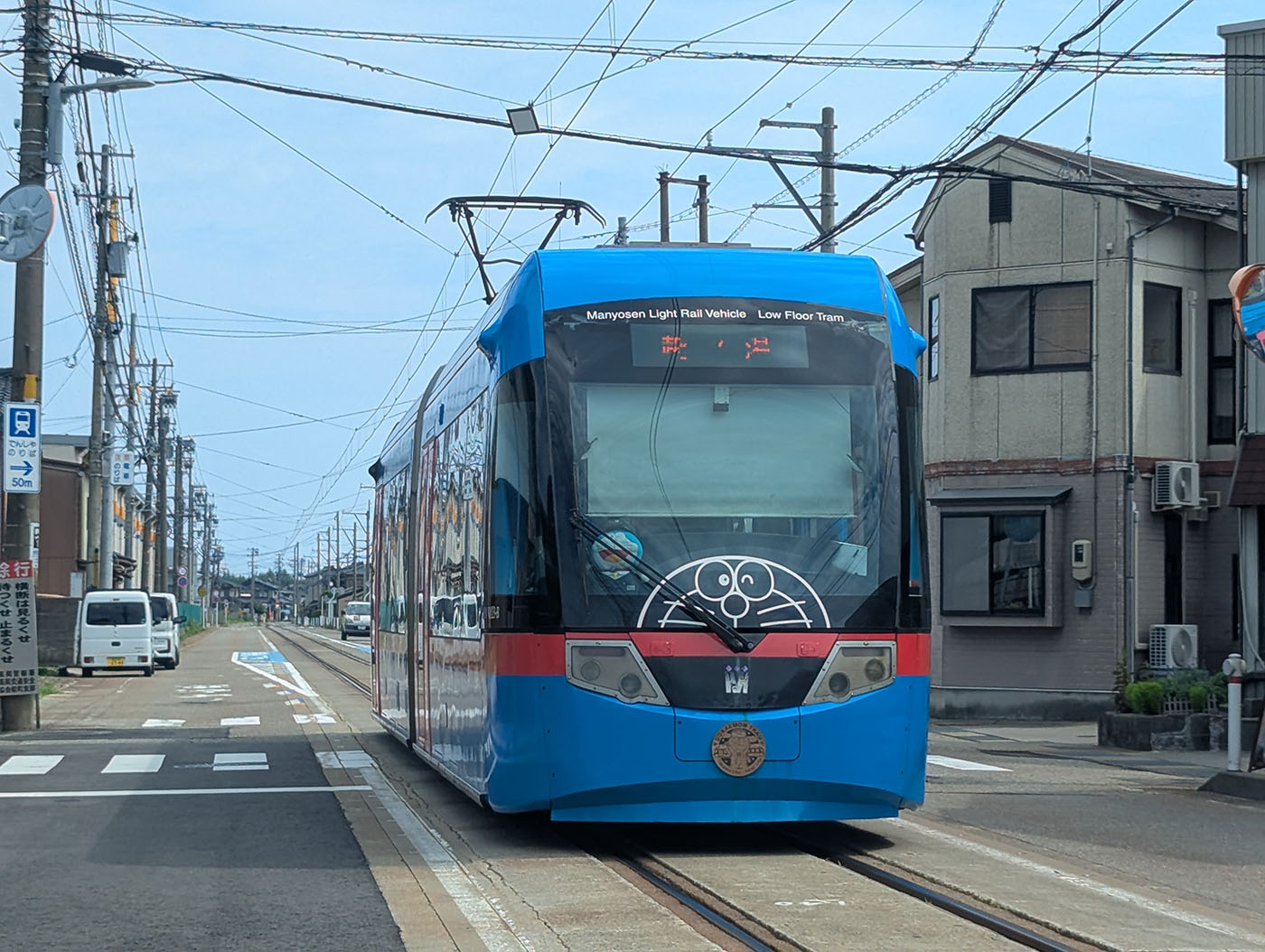 富山県の路面を走る万葉線の青い低床車両。正面に藤子・F・不二雄氏のキャラクター・ドラえもんのイラストが描かれ、「Manyosen Light Rail Vehicle Low Floor Tram」の英語表記がある。住宅街の中を路面電車が走る風景。