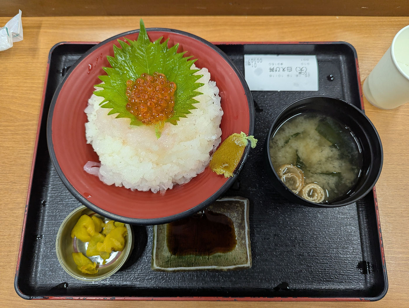 きときと食堂の白えび丼定食。赤い丼に盛られた白米の上に、透明感のある白えびがたっぷりと敷き詰められ、中央にオレンジ色のイクラと緑の大葉が添えられている。味噌汁、たくあん、醤油が付いた黒いお盆に載せられている。