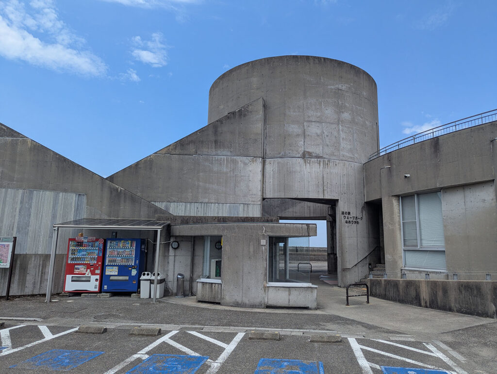 道の駅ウェーブパークなめりかわの建物外観。打ちっぱなしコンクリートの円筒形の建物が特徴的で、「道の駅 ウェーブパークなめりかわ」の看板が掲げられている。駐車場には自動販売機が設置されている。